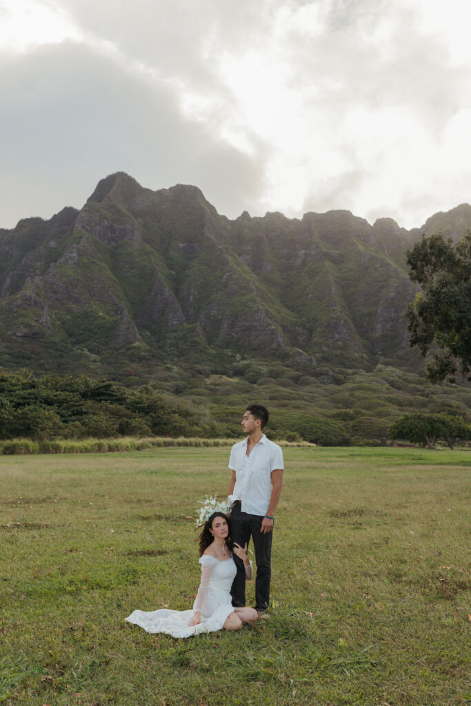 Bride and groom standing together in the foreground with Kualoa Ranch’s lush landscape and mountains in the background, captured from a distance.